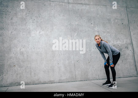 A piena lunghezza shot del giovane bella donna appoggiato su di uno sfondo grigio durante il riposo dopo ogni giorno di formazione. Donna atleta runner prendendo una pausa con Foto Stock
