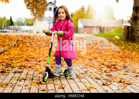 Ragazza in cappotto rosa è in sella scooter su foglie di acero. Bambino attivo nel perseguimento dello sport è di esercitare nella natura in autunno con temperature fredde ou Foto Stock