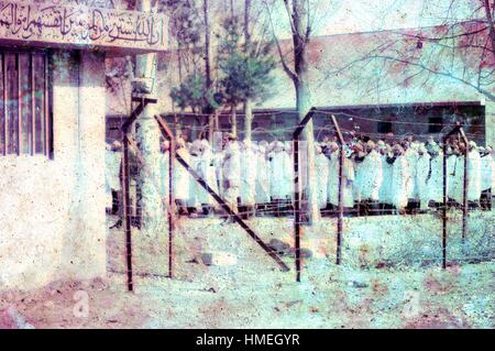 Vista attraverso reticolati di filo spinato di un gruppo di uomini vestiti in bianche vesti e lacci, in piedi con i pugni sollevata in un ampio cortile in Iran, marzo 1983. Un segno parzialmente visibile in alto a sinistra viene visualizzata in primo piano un proverbio scritto in arabo dal Al Tawbah P-111 passaggio del Corano. La porzione visibile si traduce in: 'effettivamente i credenti sono scambiati le loro vite e i loro beni al dio per [un pezzo di] il cielo..'. Danni estesi su foto originale. (Foto dalla collezione di Morse/Gado/Getty Images). Foto Stock