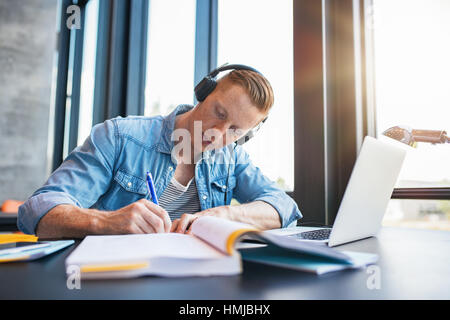 Colpo di giovane uomo che studiano in college library seduta a tavola con laptop e libri. Maschio studente universitario note di preparazione per l'esame alla libreria. Foto Stock