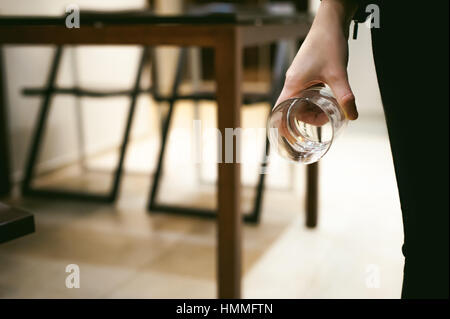 Donna bionda in cucina tenendo un bicchiere di vetro, versare l'acqua dal bollitore per soddisfare la loro sete Foto Stock