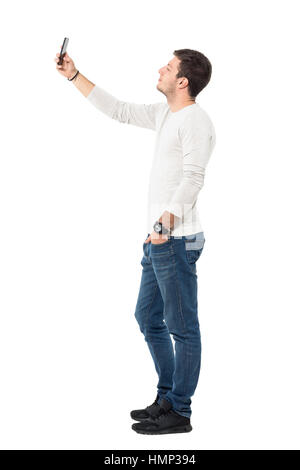 Vista laterale del giovane uomo bello indossare grigio chiaro shirt tenendo selfie con il telefono cellulare. Corpo pieno lunghezza ritratto isolate su sfondo bianco Foto Stock