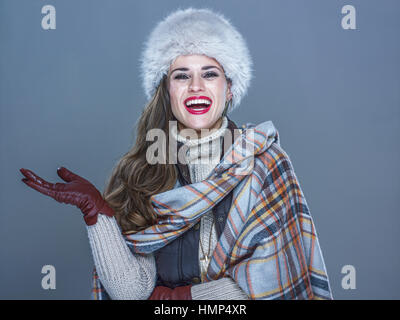 Cose d'inverno. Ritratto di sorridente donna moderna nel cappello di pelliccia isolato a freddo su sfondo blu che mostra qualcosa sulla mano vuota Foto Stock