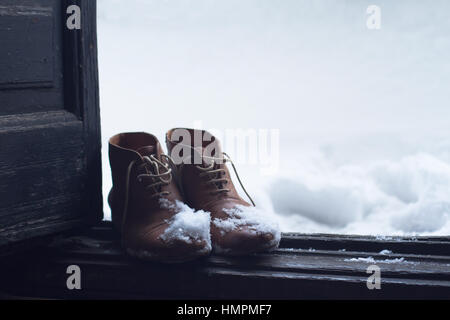 Vista frontale di una coppia di pelle marrone vintage scarpe dalla casa di legno aperto della porta d'ingresso coperto di neve in inverno Foto Stock