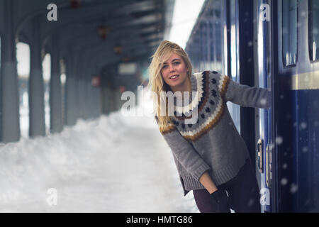 Vista frontale del biondo caucasico bella giovane donna con i capelli lunghi e un maglione pesante sorridendo vicino alla porta di un treno passeggeri in una stazione ferroviaria, Foto Stock