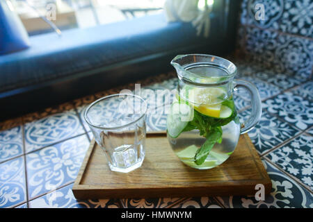 Decanter in vetro con una bevanda rinfrescante dal cetriolo, limone, menta e vetro vuoto coppa sfaccettato sorge su piastrelle con motivi in cafe. Concetto di refreshin Foto Stock