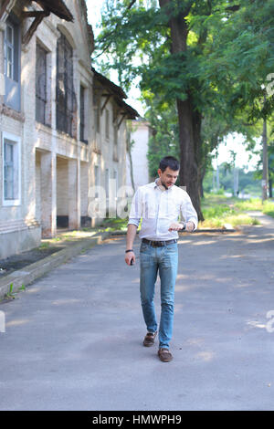 Barbuto giovani uomini uomo ragazzo va, camminando lungo la strada e cercando di guardare sul braccio. Giovane uomo vestito con una camicia bianca con striscia rossa, jeans blu e marrone Foto Stock