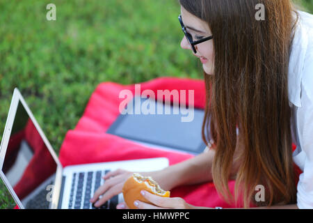 Femmina nel parco lavora dietro laptop. bellissima ragazza con gli occhiali di mangiare fast food in natura e sorridente. Foto Stock