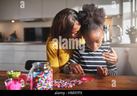 Orgoglioso giovane africana genitore femmina abbracciando carino bambino in camicia a righe rendendo craft perline forma di cuore sul tavolo in cucina Foto Stock