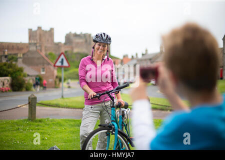 Madre che posano per una foto sulla sua moto. Un villaggio con un castello può essere visto in background, indossa un casco e abbigliamento casual. Foto Stock