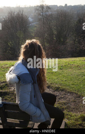 Giovane donna seduta su una panchina, torna alla fotocamera. In un parco di spazio aperto. Foto Stock