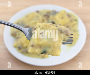 Una forcella con un pezzo di ravioli al formaggio e salsa di funghi con il pasto in background su una tavola di legno. Foto Stock