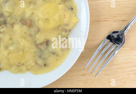 Top vista ravvicinata di un ravioli coperti con un formaggio e salsa di funghi pasto su una piastra con una forcella per il lato su una tavola di legno alto. Foto Stock