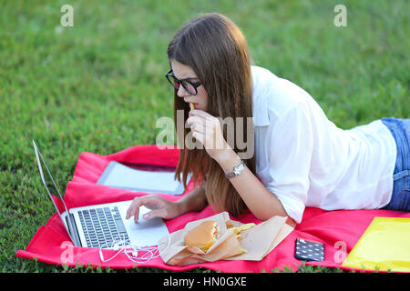 Femmina nel parco lavora dietro laptop. bellissima ragazza con gli occhiali di mangiare fast food in natura e sorridente. Foto Stock