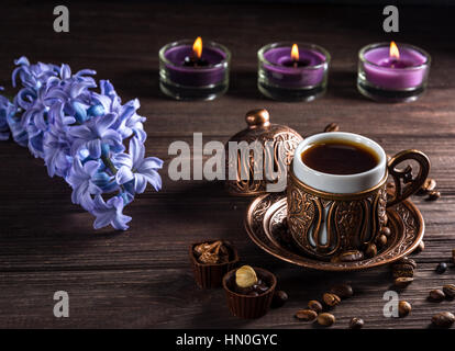 Tazza di caffè nero, candele e fiori su un sfondo di legno. Tasto scuro. Foto Stock