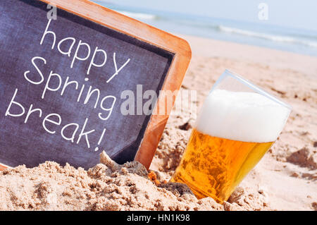 Primo piano di un di legno-incorniciato lavagna con il testo happy spring break scritto in esso e un bicchiere di birra rinfrescante, sulla sabbia di una spiaggia Foto Stock