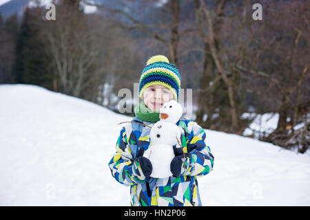 Carino bambino, edificio pupazzo di neve e giocare con essa sulla cima della montagna, inverno Foto Stock