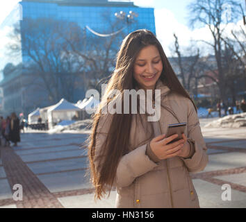 Stupenda bella bruna giovane donna di messaggistica sul smart-phone alla strada di città sullo sfondo. Pretty girl avente smart phone conversazione in sun Foto Stock