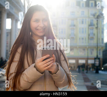 Stupenda bella bruna giovane donna di messaggistica sul smart-phone alla strada di città sullo sfondo. Pretty girl avente smart phone conversazione in sun Foto Stock