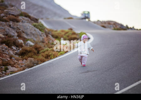 Felice bambina correndo giù per strada. genitori' auto parcheggiate lungo la strada in distanza. Foto Stock