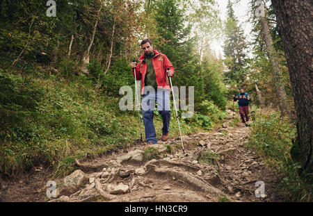 Due giovani uomini che indossano gli zaini e il trasporto di pali trekking a piedi giù per un sentiero forestale durante le escursioni nel deserto Foto Stock