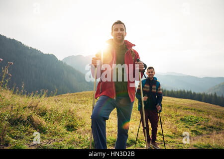 Ritratto di due giovani uomini che indossano gli zaini e il trasporto di pali trekking in piedi in un campo durante le escursioni in collina Foto Stock