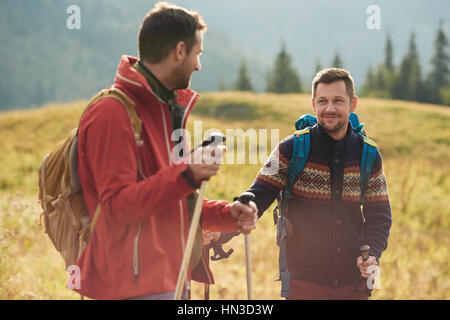 Due giovani uomini che indossano gli zaini e il trasporto di pali trekking parlare insieme in un campo durante le escursioni in collina Foto Stock