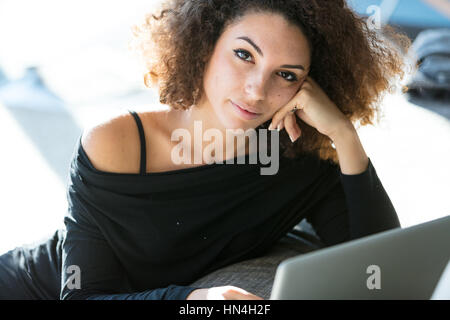 Malinconici attraente giovane donna che guarda la telecamera come lei si rilassa sotto il sole su un divano a casa con il suo computer portatile sul suo giro Foto Stock