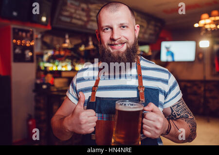 Barman Barbuto con tatuaggi di indossare un grembiule in piedi al bar e in mano un bicchiere di birra Foto Stock