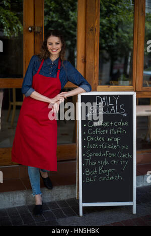 Ritratto di sorridente cameriera appoggiata sulla scheda menu al di fuori del cafe Foto Stock