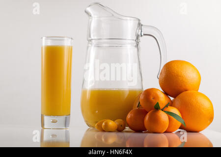 Composizione del bicchiere pieno di succo di arancia, poche arance e mandarini e un recipiente su una superficie bianca Foto Stock