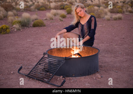 Ragazza bionda seduta da una buca per il fuoco con vento di notte con la fiamma nel deserto Foto Stock