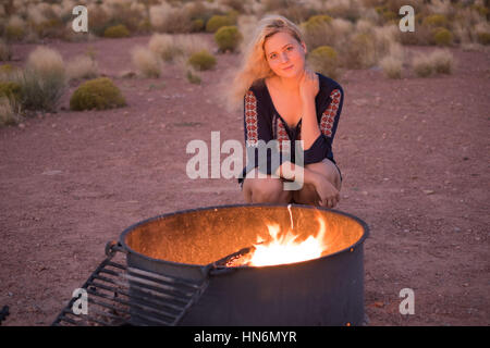 Ragazza bionda seduta da una buca per il fuoco con vento di notte con la fiamma nel deserto Foto Stock