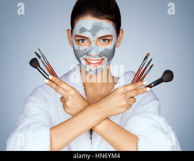 Ragazza allegra con set di spazzole. Foto giovane donna con maschera di argilla su sfondo blu. Beauty & Skin care concept Foto Stock