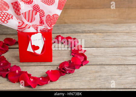 Confezione regalo circondato con il rosso dei petali di rosa luogo in forma di cuore su sfondo di legno Foto Stock