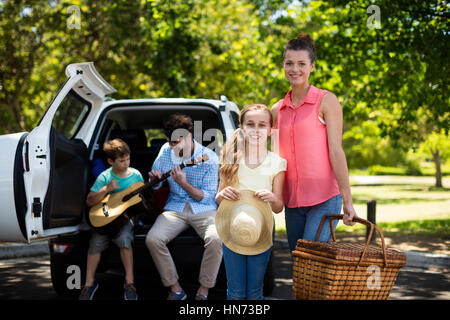 Ritratto di Madre e figlia in piedi con Cesto picnic mentre il padre e il figlio a suonare la chitarra in background in posizione di parcheggio Foto Stock