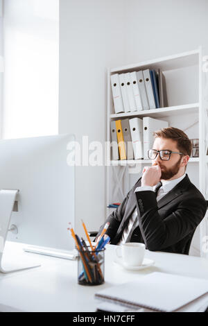 Foto di attraente giovane imprenditore barbuto seduto in ufficio mentre si utilizza il computer. Foto Stock
