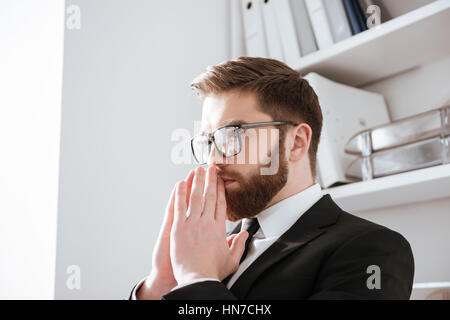 Foto di attraente giovane imprenditore barbuto seduto in ufficio mentre guardando a parte. Foto Stock