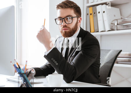 Foto di grave giovane imprenditore barbuto seduto in ufficio mentre guardando la fotocamera e tenendo una matita. Foto Stock