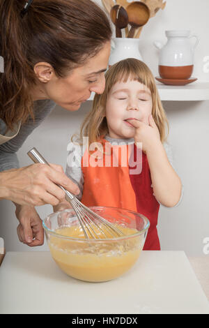 Tre anni bambino succhiare il suo dito per il gusto di panna montata, accanto alla donna, nel lavoro di squadra, la realizzazione e la cottura di una torta di pan di spagna in cucina home Foto Stock