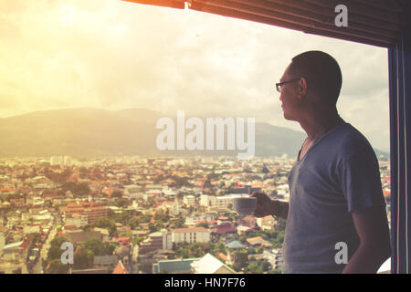 Uomo asiatico holding tazza di caffè in ufficio egli a guardare fuori dalla finestra. Cityscape sfondo. Messa a fuoco selettiva e soft filtro flare. Foto Stock