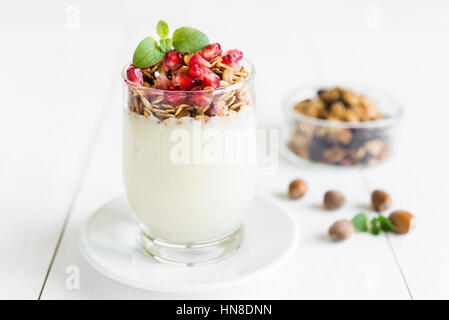 Colazione sana nozione. Crema alla vaniglia con muesli fresco in un ripiano in vetro con melograno e menta disposti su bianco tavolo in legno. Foto Stock