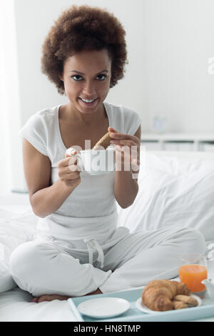 Giovane africano donna americana avente una rilassante colazione a letto, ella è immergendo i cookie in una tazza di tè Foto Stock