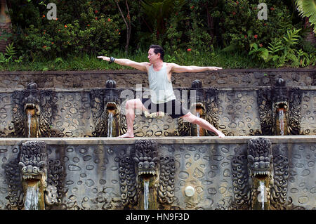 Middle-Aged Uomo asiatico detiene un guerriero di Hatha Yoga pone a Banjar Hot Springs per piscine di Bali, Indonesia come acqua si riversa da creature Dragon-Like' bocche Foto Stock