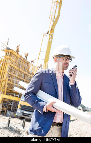 Supervisore utilizzando un walkie-talkie tenendo blueprint al sito in costruzione Foto Stock