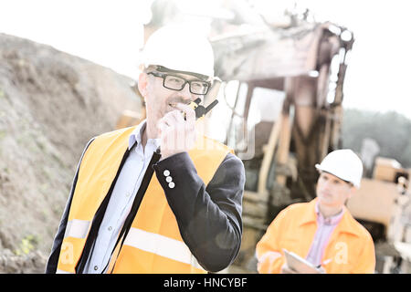 Ingegnere felice utilizzando un walkie-talkie sul sito di costruzione con il collega in background Foto Stock