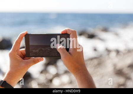 Uomo di fotografare il mare stando in piedi sul litorale giorno Foto Stock