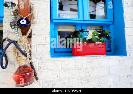 Vecchio hookah-shisha-narghile-waterpipe con nazar-cordone blu amuleto appeso al muro di calcestruzzo di una casa con finestra blu e rosso plantpot-street o Foto Stock