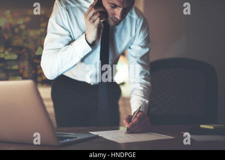 Imprenditore su telefono firma documento in ufficio durante la notte in piedi alla scrivania Foto Stock