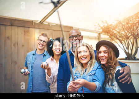 Cinque amici giovani uomini e donne che assumono selfie con selfie stick holding petanque sfere metalliche in mani, sorridente e guardando in alto Foto Stock